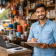 Smiling Indian small business owner standing in a roadside hardware shop with a clipboard and laptop, representing grassroots entrepreneurship in India.